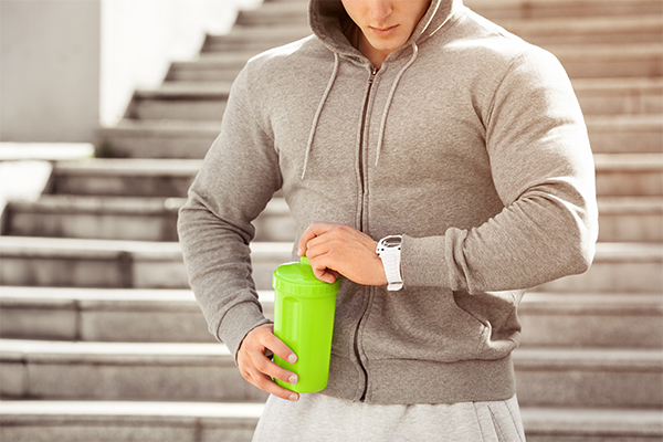 man in hoodie holding shaker bottle of whey protein looking at watch in front of stairs