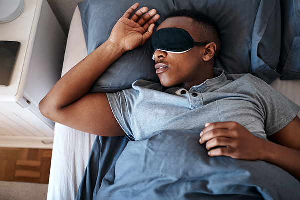 Man asleep under weighted blanket wearing eye mask.