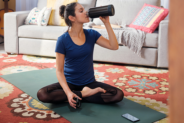 Woman on yoga mat in living room drinking water.